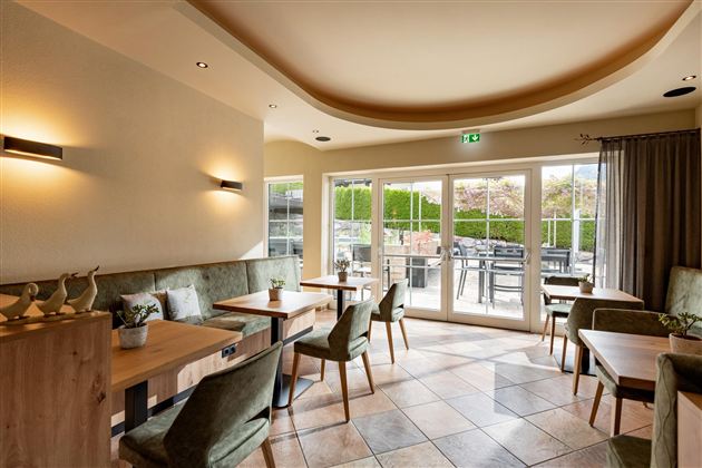 Ein gemütliches Café mit Holztischen und grünen Stühlen. Große Fenster bieten einen Blick auf den Außenbereich.