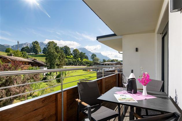 Ein schöner Balkon mit Blick auf grüne Wiesen und Berge. Ein Tisch mit Blumen und einer Flasche Wein lädt zum Entspannen ein.