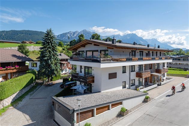 Ein modernes Gebäude in einer schönen Landschaft mit Bergen im Hintergrund. Die Straße vor dem Haus ist ruhig und von Bäumen gesäumt.