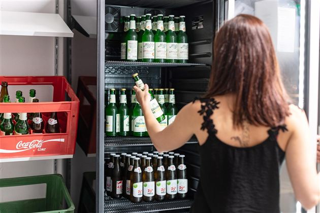 Eine Person steht vor einem Kühlschrank mit verschiedenen Bierflaschen. Im Hintergrund sind leere Kisten zu sehen.