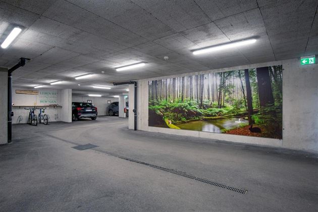 Eine moderne Tiefgarage mit einem großen Wandbild eines Waldes. Im Hintergrund stehen mehrere Fahrzeuge und Fahrräder sind abgestellt.