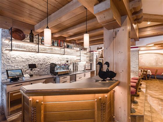 Eine gemütliche Bar mit Holzelementen und eleganter Beleuchtung. Im Hintergrund sind Getränke und Sitzmöglichkeiten sichtbar.