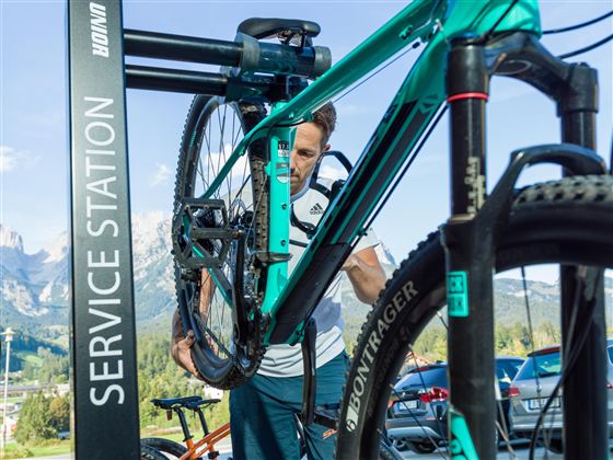 A person is working on a bicycle at a service station. In the background, mountains and cars can be seen.
