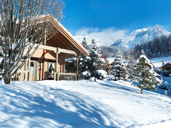Ein gemütliches Holzhaus im Schnee, umgeben von verschneiten Bäumen. Im Hintergrund sind schneebedeckte Berge und ein blauer Himmel zu sehen.