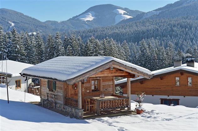 A cozy wooden cabin in the snow, surrounded by snow-covered mountains and forests. The clear sky and winter landscape create a peaceful atmosphere.
