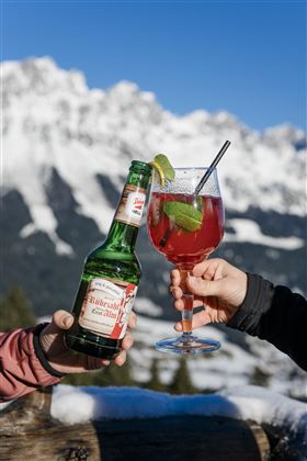 Ein Bier und ein Cocktail werden in den Bergen angestoßen. Im Hintergrund sind schneebedeckte Berge und blauer Himmel zu sehen.