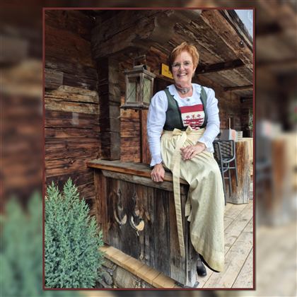 A smiling woman is sitting on a wooden rock in front of a rustic cabin. She is wearing a traditional outfit and has short, reddish-brown hair.