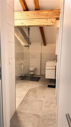 Een moderne badkamer met lichte tegels en houten balken. Op de achtergrond zijn een douche en een wastafel te zien.