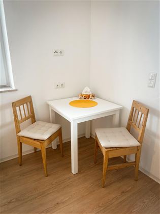 Een kleine eetruimte met een witte tafel en twee houten stoelen. Op de tafel ligt een oranje placemat.