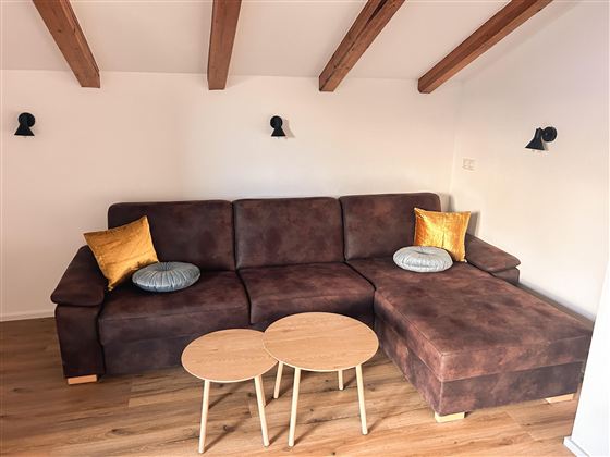 Een gezellige zithoek met een bruin bank en decoratieve kussens. Daar horen ook twee kleine tafeltjes van hout bij, alles in een lichte, moderne ruimte.