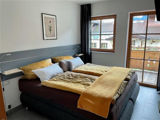 Ein gemütliches Schlafzimmer mit einem großen Bett und warmen Farbtönen. Es gibt ein Fenster mit Blick auf einen Balkon und die umliegenden Gebäude.