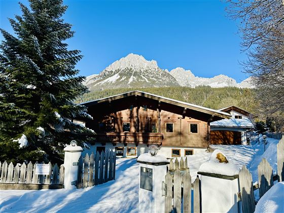 A cozy wooden house in the snow with snow-covered mountains in the background. The surroundings are characterized by fir trees and a white fence.