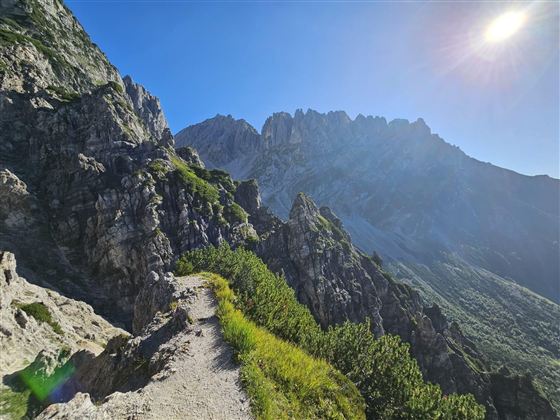 Een schilderachtig wandelpad leidt door rotsachtige bergen onder een heldere blauwe lucht. De zon straalt helder over het landschap.