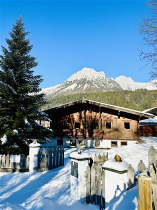 A cozy wooden house in the snow with a wooden protective wall. In the background, majestic mountains rise up under a clear blue sky.