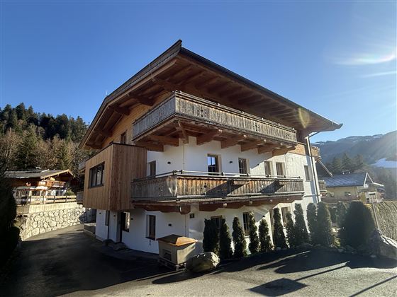 Ein modernes Haus im alpinen Stil mit Holzverkleidung und mehreren Balkonen. Die Umgebung ist von grünen Bäumen und Bergen umgeben.