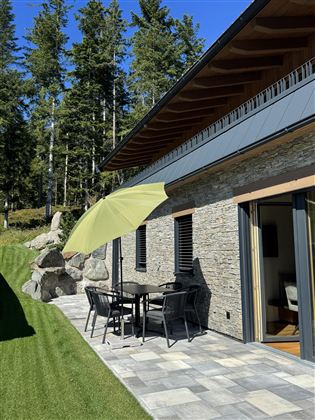 Een terras met een tafel en stoelen, overdekt door een groene parasol. Op de achtergrond zijn bomen en een gezellig huis te zien.