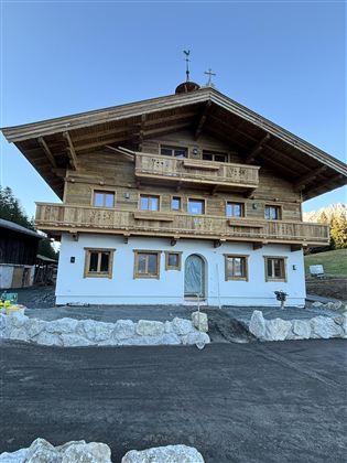 A traditional wooden house with a balcony in alpine style. The façade is partly painted white, and there is a well-kept outdoor area.