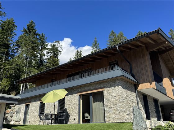 Een modern huis met een steen- en houten gevel, omringd door bomen. Op het terras staan tafels en stoelen, evenals een gele parasol.