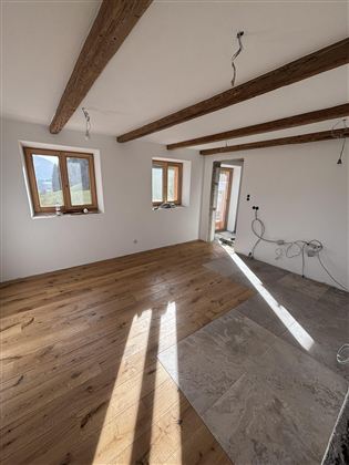 A bright room with wooden flooring and exposed wooden beams. The walls are still incomplete and there are windows that let in a lot of light.