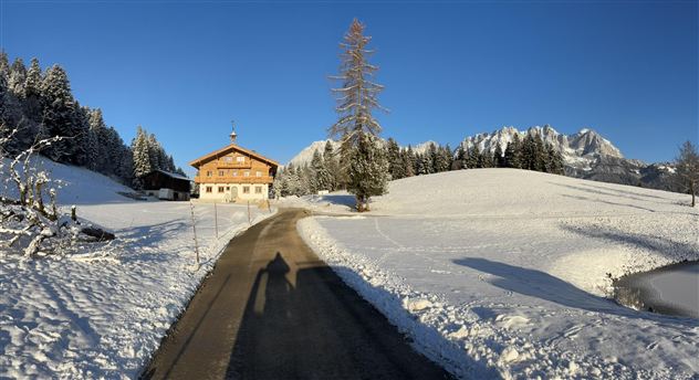 A snowy landscape with a large wooden house and snow-covered mountains in the background. The path leads through the idyllic winter scenery.