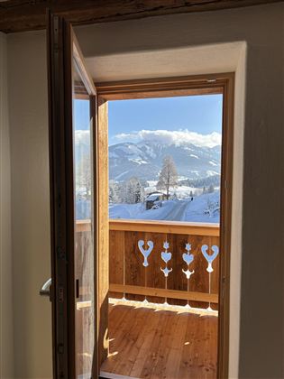 An open window with a view of snow-covered mountains and a wooden balcony. The view is clear and sunny.