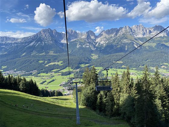 Een schilderachtig berglandschap met groene weilanden en hoge bergen op de achtergrond. Een kabelbaan rijdt door de mooie natuur.