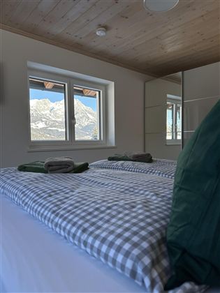 A cozy bedroom with a large window and a view of the mountains. The beds are decorated with checkered bedding and green cushions.