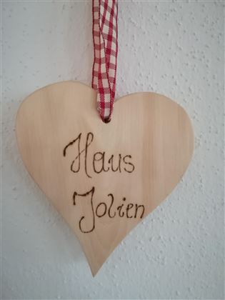 A heart-shaped wooden plaque with the inscription "Haus Jolien". It is equipped with a red checked ribbon for hanging.
