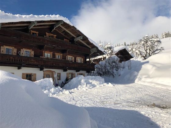 A cozy wooden house in a snowy landscape. The snow is high and the sky is clear and blue.