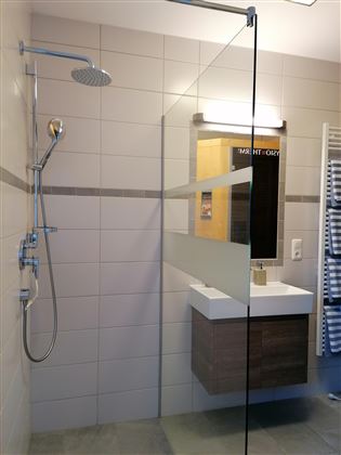 Een moderne badkamer met een glazen douche en een eenvoudige wastafel. De muren zijn licht betegeld en de verlichting zorgt voor een aangename sfeer.