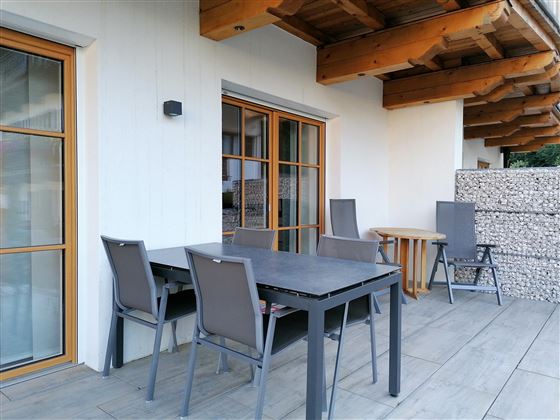Een moderne terras met een tafel en stoelen. De muren zijn licht en de overkapping biedt bescherming tegen zon en regen.