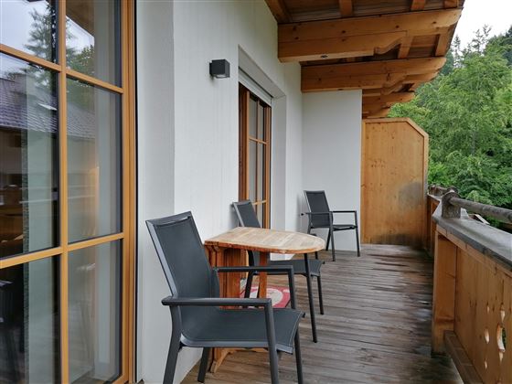 Ein gemütlicher Balkon mit einem Holztisch und zwei Stühlen. Im Hintergrund sind grüne Bäume sichtbar.