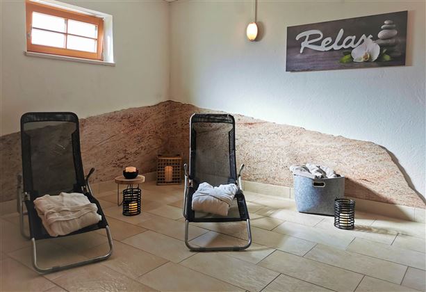 Ein ruhiger Entspannungsraum mit zwei Liegestühlen und sanfter Beleuchtung. Auf dem Boden liegen Handtücher, und an der Wand hängt ein Schild mit der Aufschrift "Relax".