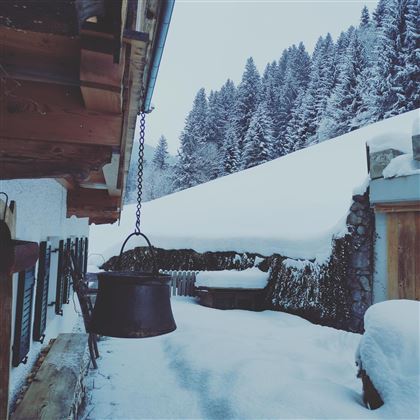 A snowy winter landscape with a wooden house and dense, snow-covered trees in the background. A bell chimes hangs on the side of the house.