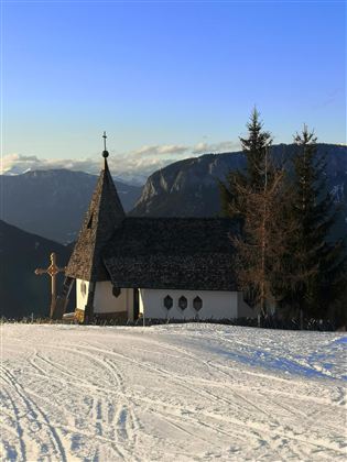A small church in a snowy landscape with mountains in the background. The sky is clear and blue.