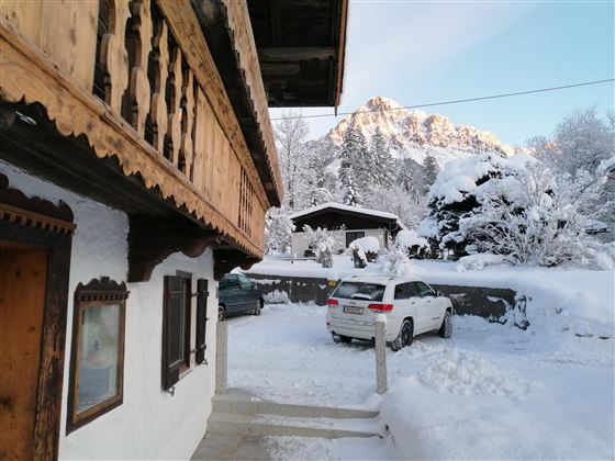 A winter landscape with snow-covered trees and mountains in the background. A traditional wooden house is visible in the foreground, next to a car parked in the snow.
