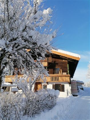 A cozy wooden house in the snow under a clear blue sky. Surrounding the house are snow-covered trees and a snow-covered pathway.
