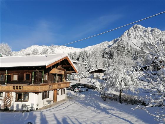A cozy wooden house in a snowy landscape. In the background, a majestic mountain range rises under a clear blue sky.