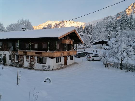 A charming, wintry farmhouse surrounded by snow. The mountains in the background are coated with snow, and the atmosphere is calm and picturesque.