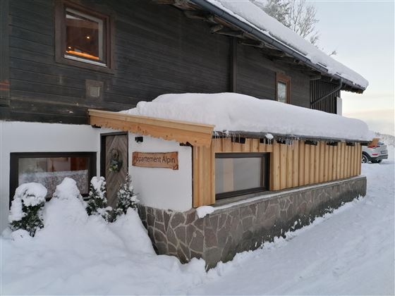 A cozy wooden house with a snow-covered roof in a wintry landscape. The entrance area is simply and invitingly designed.