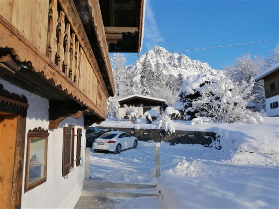A traditional wooden house in the snow, surrounded by a wintry landscape. In the background, snow-covered mountains and a clear blue sky can be seen.