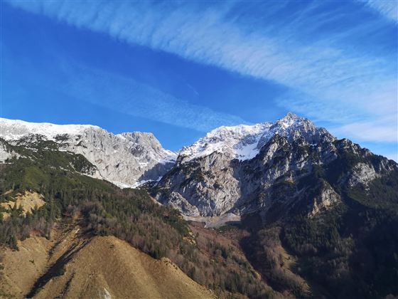 An impressive mountain landscape with snow-covered peaks and a bright blue sky. In the foreground, green hills and forests are visible.