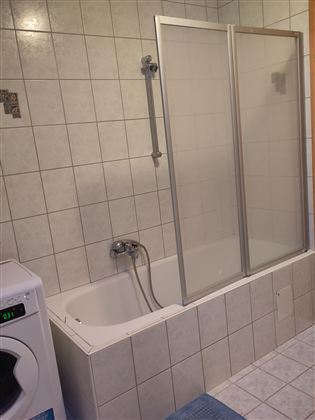 Een eenvoudige badkamer met een bad en een eigen douchewand. Aan de linkerkant staat een wasmachine.