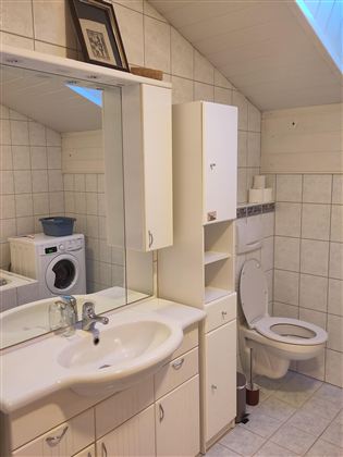 Een moderne badkamer met een wasmachine, een wastafel en een toilet. De wanden zijn betegeld met lichte tegels en een raam zorgt voor daglicht.