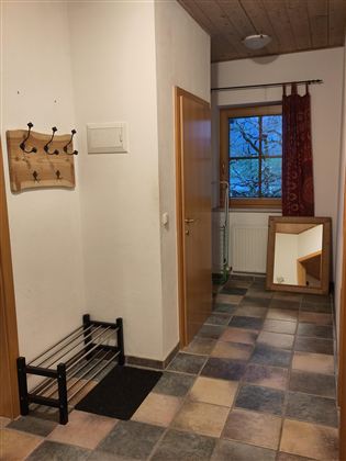 Een smalle gang met een hangende kapstok en een spiegel. Het licht valt door een raam dat omrand is met een rode gordijn.