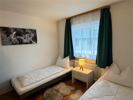 Ein einfaches Zimmer mit zwei Einzelbetten und einem Nachttisch. Die Vorhänge sind grün und es gibt ein warmes Licht von einer Lampe.