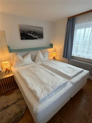 Ein gemütliches Schlafzimmer mit einem Doppelbett und zwei Nachttischen. Große Fenster sorgen für viel Tageslicht und eine entspannte Atmosphäre.