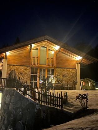 Ein gemütliches Holzhaus in der Nacht, beleuchtet von warmem Licht. Es steht in einer verschneiten Umgebung mit einem kleinen Garten und einem Zaun.