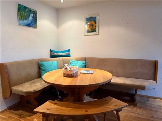 Een gezellige eetruimte met een ronde houten tafel en een bank. De muren zijn versierd met foto's, en er zijn kleurrijke kussens op de bank.