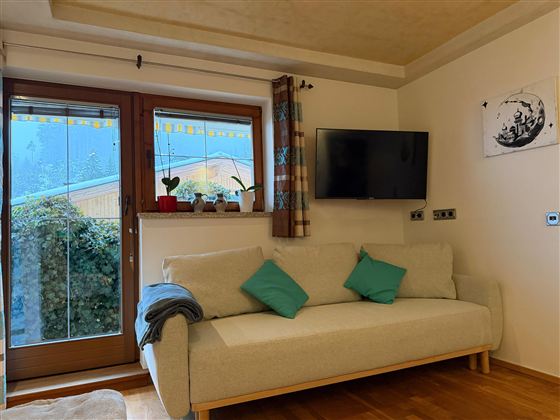 Ein gemütliches Wohnzimmer mit einem hellen Sofa und grünen Kissen. Große Fenster bieten einen Blick auf Pflanzen im Freien und einen Fernseher an der Wand.